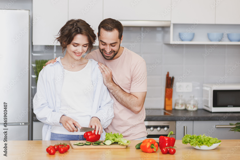 Cheerful young couple two friends guy girl in casual clothes preparing vegetable salad cooking food in light kitchen at home. Dieting family healthy lifestyle concept. Mock up copy space. Hugging.