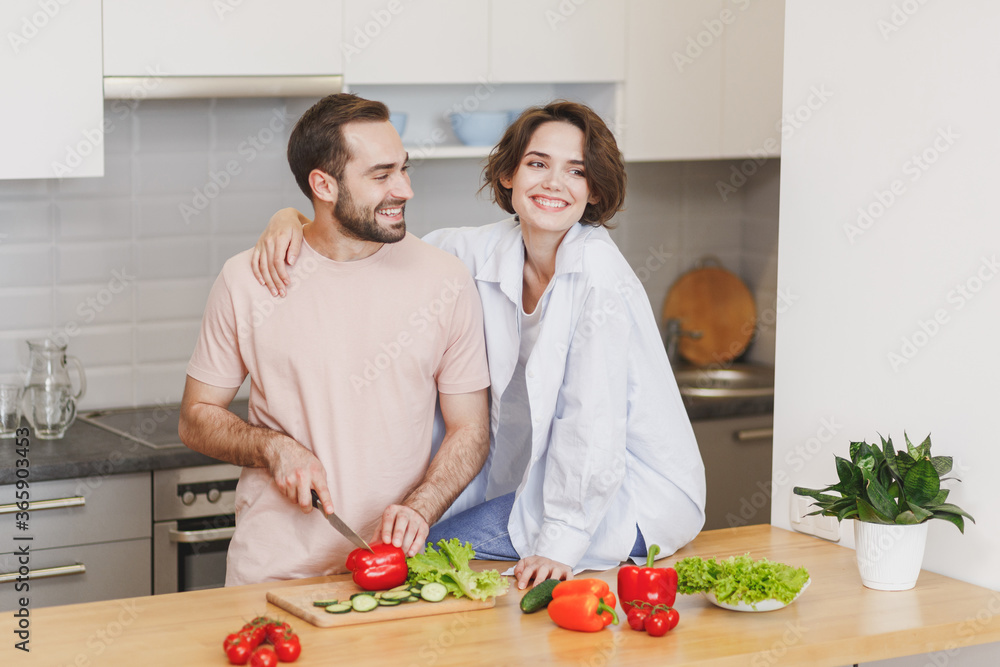Smiling young couple two friends guy girl in casual clothes hugging sit on table preparing vegetable salad cooking food in light kitchen at home. Dieting healthy lifestyle concept. Mock up copy space. Smiling young couple two friends guy girl in casual clothes hugging sit on table preparing vegetable salad cooking food in light kitchen at home. Dieting healthy lifestyle concept. Mock up copy space.