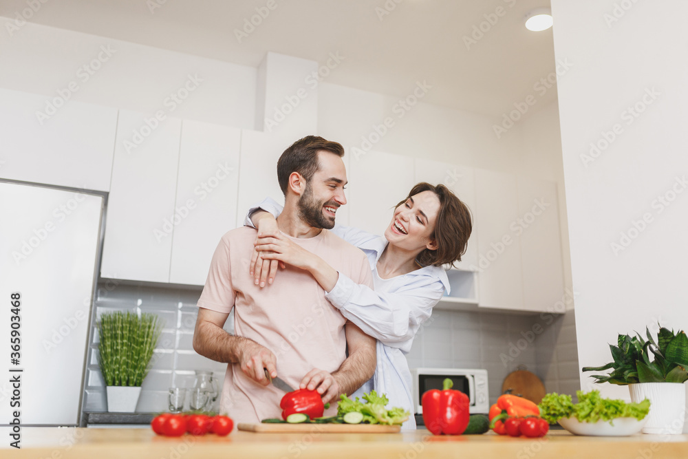 Laughing young couple two friends guy girl in casual clothes preparing vegetable salad cooking food in light kitchen at home. Dieting family healthy lifestyle concept. Mock up copy space. Hugging. - obrazy, fototapety, plakaty 