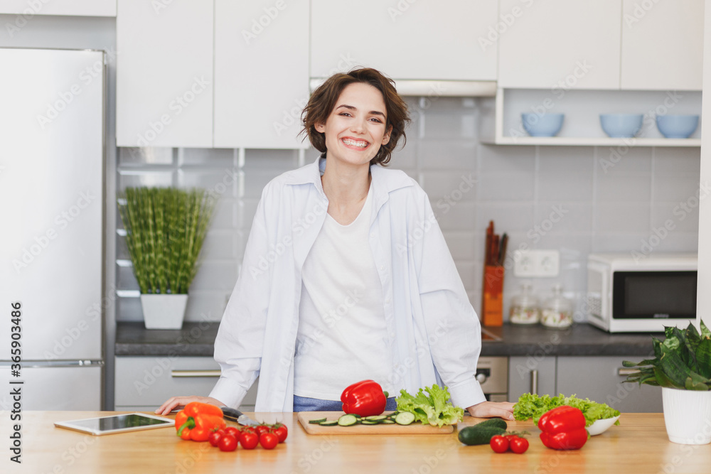 Smiling cheerful young brunette housewife woman girl in white casual clothes preparing vegetable salad cooking food in light kitchen at home. Dieting healthy lifestyle concept. Mock up copy space. Smiling cheerful young brunette housewife woman girl in white casual clothes preparing vegetable salad cooking food in light kitchen at home. Dieting healthy lifestyle concept. Mock up copy space.