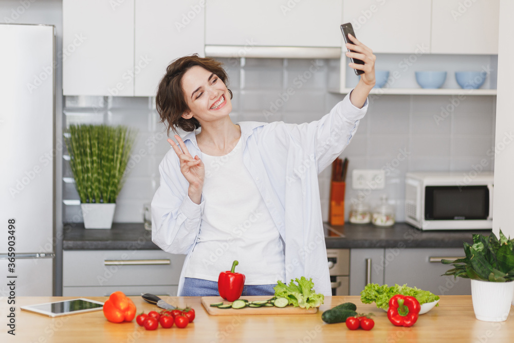 Smiling housewife woman in casual clothes preparing vegetable salad cooking food in light kitchen at home. Dieting healthy lifestyle concept. Doing selfie shot on mobile phone, showing victory sign. Smiling housewife woman in casual clothes preparing vegetable salad cooking food in light kitchen at home. Dieting healthy lifestyle concept. Doing selfie shot on mobile phone, showing victory sign.