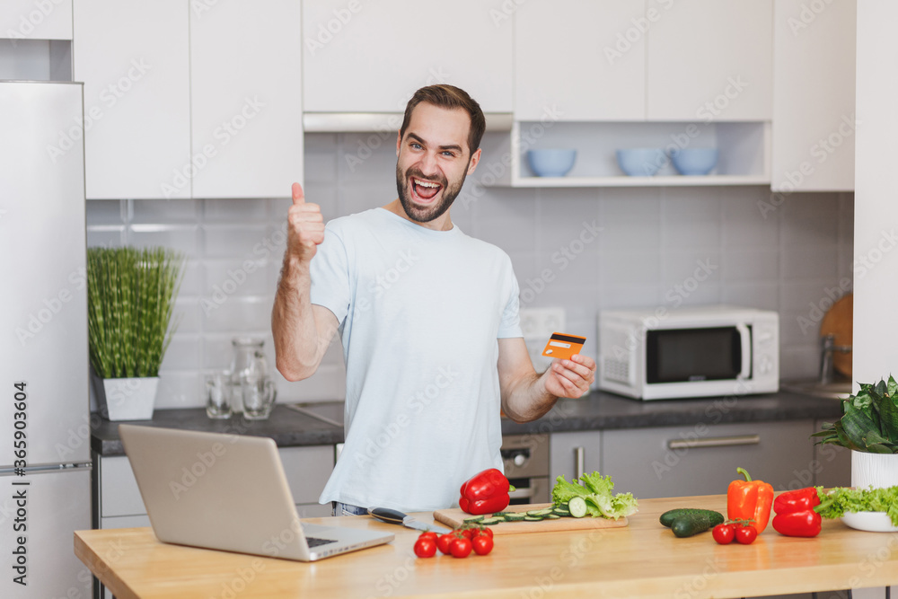 Joyful young man guy in white t-shirt using laptop computer hold credit bank card showing thumb up preparing vegetable salad cooking food in light kitchen at home. Dieting healthy lifestyle concept.