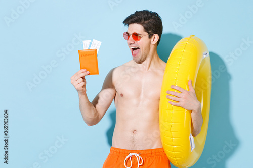 Excited young man guy in eyeglasses isolated on pastel blue wall background studio portrait. People summer vacation rest lifestyle concept. Mock up copy space. Hold inflatable ring passport tickets.