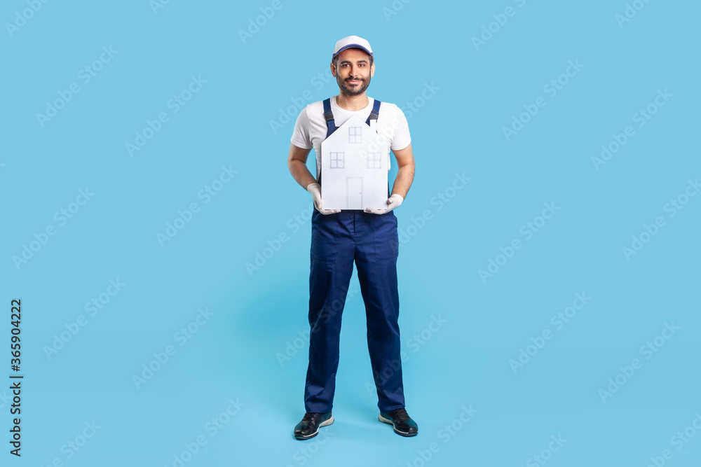 Full length handyman, building worker in uniform holding paper house. Mover guy offering help in relocation, home repair services and maintenance. indoor studio shot isolated on blue background Full length handyman, building worker in uniform holding paper house. Mover guy offering help in relocation, home repair services and maintenance. indoor studio shot isolated on blue background