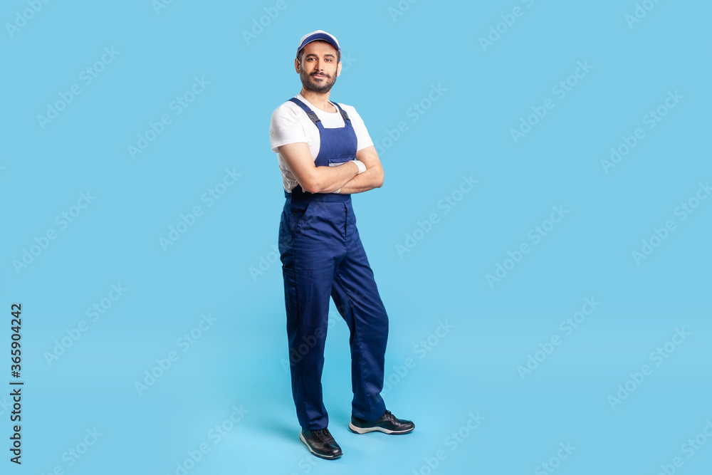 Full length confident professional handyman in overalls standing with crossed hands, smiling at camera. Profession of service industry, house repair. indoor studio shot isolated on blue background Full length confident professional handyman in overalls standing with crossed hands, smiling at camera. Profession of service industry, house repair. indoor studio shot isolated on blue background
