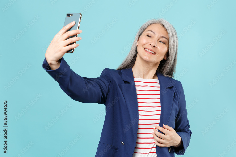 Smiling gray-haired business woman in blue suit posing isolated on pastel blue background studio. Achievement career wealth business concept. Mock up copy space. Doing selfie shot on mobile phone. Smiling gray-haired business woman in blue suit posing isolated on pastel blue background studio. Achievement career wealth business concept. Mock up copy space. Doing selfie shot on mobile phone.