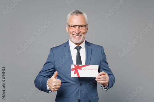 Smiling elderly gray-haired business man in classic blue suit shirt tie isolated on grey background studio portrait. Achievement career wealth business concept. Hold gift certificate showing thumb up.