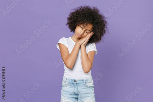 Little african american kid girl 12-13 years old in white t-shirt isolated on violet background studio portrait. Childhood lifestyle concept. Mock up copy space. Sleep with folded hands under cheek.
