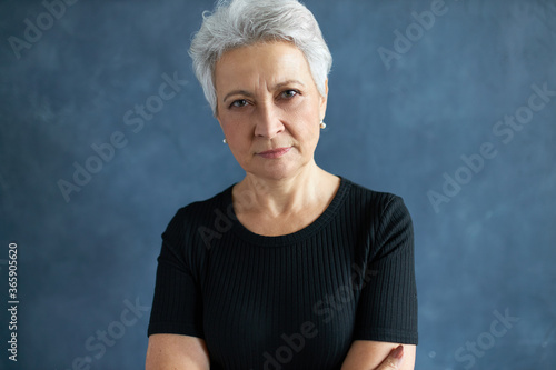 Human emotions and feelings. Headshot of casually dressed 60 year old retired female with gray hair keeping arms crossed on chest, staring at camera with scrutinizing suspicious look, narrowing eyes