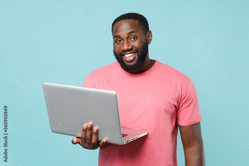 Smiling young african american man guy in casual pink t-shirt isolated on pastel blue wall background studio portrait. People lifestyle concept. Mock up copy space. Working on laptop pc computer. Smiling young african american man guy in casual pink t-shirt isolated on pastel blue wall background studio portrait. People lifestyle concept. Mock up copy space. Working on laptop pc computer.