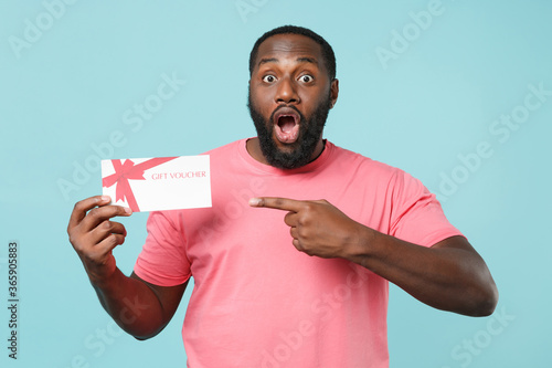 Shocked young african american man guy in casual pink t-shirt isolated on blue wall background studio portrait. People lifestyle concept. Mock up copy space. Pointing index finger on gift certificate. © ViDi Studio