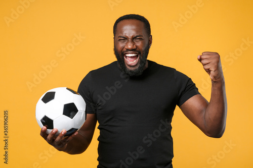 Screaming african american man guy football fan in black t-shirt isolated on yellow background. Sport family leisure lifestyle concept. Cheer up support favorite team with soccer ball, clenching fist. © ViDi Studio
