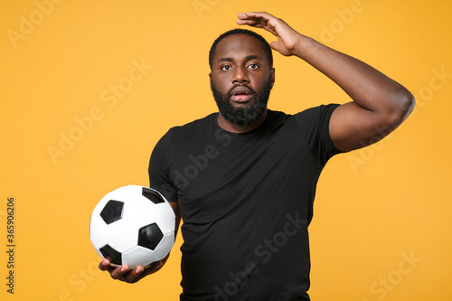 Displeased african american man football fan in black t-shirt isolated on yellow background. Sport family leisure lifestyle concept. Cheer up support favorite team with soccer ball put hand on head.