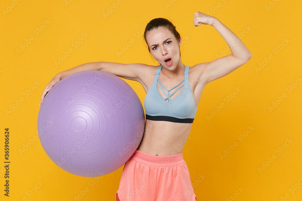 Strong young fitness sporty woman in sportswear working out isolated on yellow background studio portrait. Workout sport motivation concept. Mock up copy space. Hold fitball, showing biceps, muscles.