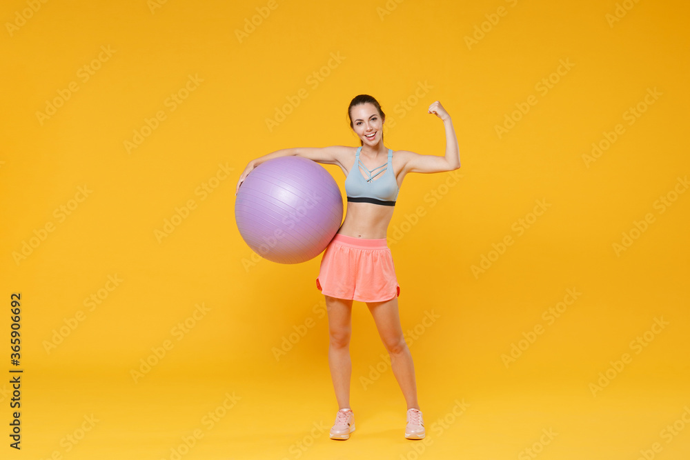 Full length portrait of smiling young fitness woman girl in sportswear working out isolated on yellow wall background. Workout sport motivation lifestyle concept. Hold fitball showing biceps, muscles. Full length portrait of smiling young fitness woman girl in sportswear working out isolated on yellow wall background. Workout sport motivation lifestyle concept. Hold fitball showing biceps, muscles.