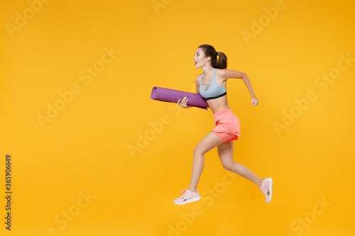 Full length portrait beautiful young fitness woman in sportswear working out isolated on yellow wall background. Workout sport motivation lifestyle concept. Mock up copy space. Jumping, hold yoga mat.