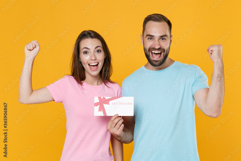 Excited young couple two friends guy girl in blue pink t-shirts posing isolated on yellow background studio. People lifestyle concept. Mock up copy space. Hold gift certificate, doing winner gesture. Excited young couple two friends guy girl in blue pink t-shirts posing isolated on yellow background studio. People lifestyle concept. Mock up copy space. Hold gift certificate, doing winner gesture.