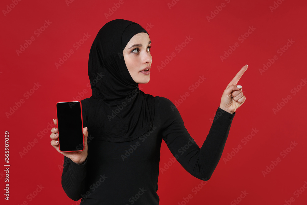 Arabian muslim woman in hijab black clothes isolated on red background. People religious lifestyle concept. Mock up copy space. Hold mobile phone with blank empty screen pointing index finger aside. Arabian muslim woman in hijab black clothes isolated on red background. People religious lifestyle concept. Mock up copy space. Hold mobile phone with blank empty screen pointing index finger aside.