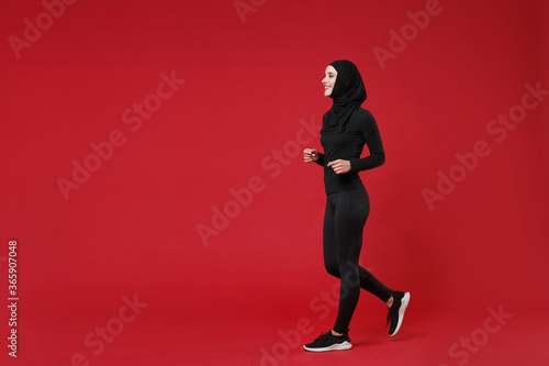 Full length portrait side view of smiling young arabian muslim woman in hijab black clothes posing isolated on red background. People religious lifestyle concept. Mock up copy space. Looking aside.