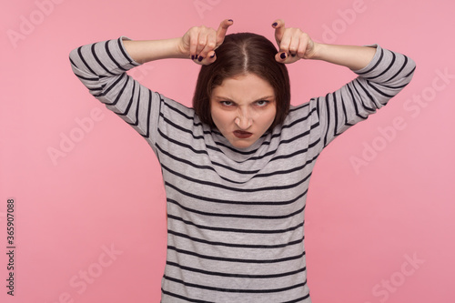 Canvas Print Portrait of funny angry woman in striped sweatshirt showing bull horn gesture and looking with hate rage at camera, threatening and provoking conflict