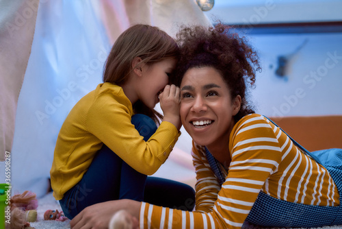 Trust and care. African american woman baby sitter entertaining caucasian cute little girl. They are gossiping and telling secrets sitting in kids room. Leisure activities, babysitting concept