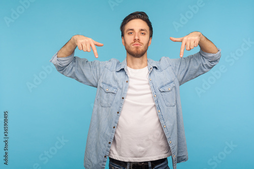 Here and right now! Portrait of bossy tyrant man in denim shirt pointing down and looking with arrogance, demanding immediate submission, demonstrating authority. indoor studio shot, blue background