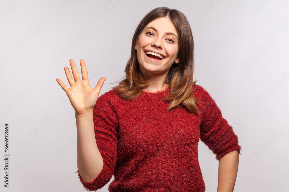 Hello, nice to meet you! Portrait of happy brunette girl in shaggy sweater raising hand waving hi and smiling friendly, looking at camera with hospitable expression. indoor studio shot isolated Hello, nice to meet you! Portrait of happy brunette girl in shaggy sweater raising hand waving hi and smiling friendly, looking at camera with hospitable expression. indoor studio shot isolated