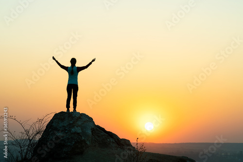 Silhouette of a woman hiker standing alone on big stone at sunset in mountains. Female tourist raising her hands up on high rock in evening nature. Tourism, traveling and healthy lifestyle concept.