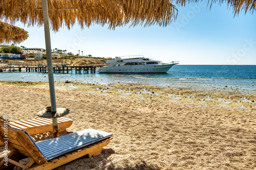 Wooden deck chairs under rough straw sun umbrella on sea beach and big white yacht ship in water near shore on sunny summer day.