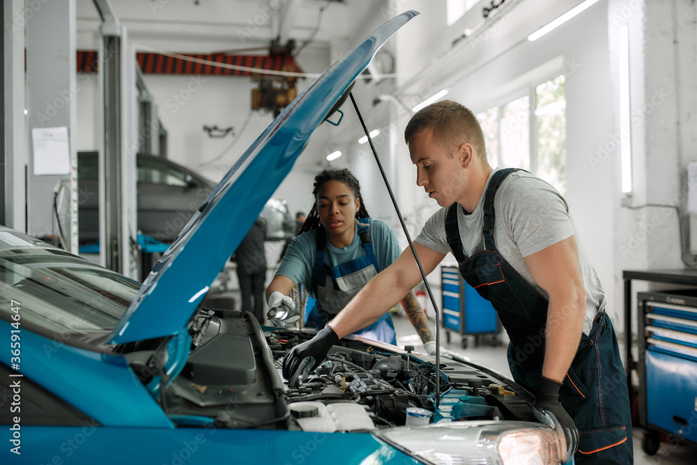 True help. Professional male mechanic repairing car engine, tighten, screw with spanner while his female colleague holding torch under car hood at service station True help. Professional male mechanic repairing car engine, tighten, screw with spanner while his female colleague holding torch under car hood at service station