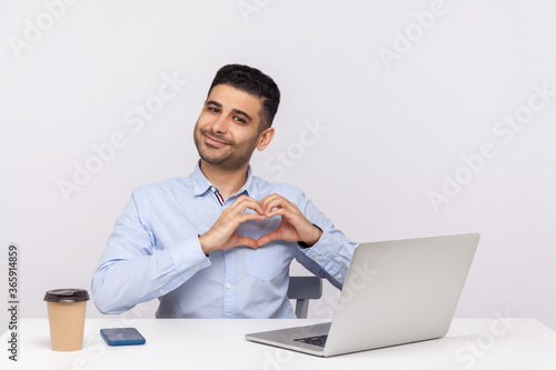 Successful happy businessman or employee sitting office workplace with laptop on desk, making heart shape gesture and smiling satisfied, loves his job. indoor studio shot isolated on white background