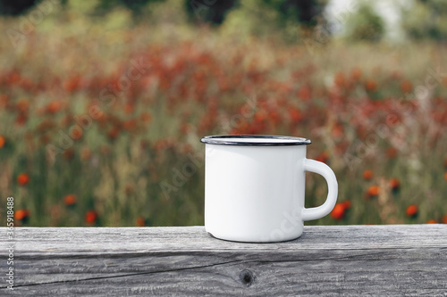 Close up of metal mug on old wooden table, board with defocused blooming mountain meadow. Outdoor tea, coffee time. Mockup of white enamel cup. Lifestyle relax, trekking and camping concept.