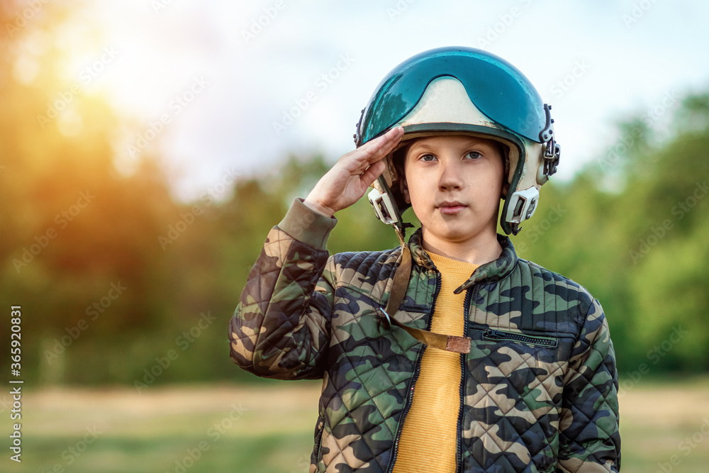 A boy in a pilot's helmet on a background of greenery salutes. Dream concept, choice of profession, military service. Copy space. A boy in a pilot's helmet on a background of greenery salutes. Dream concept, choice of profession, military service. Copy space.
