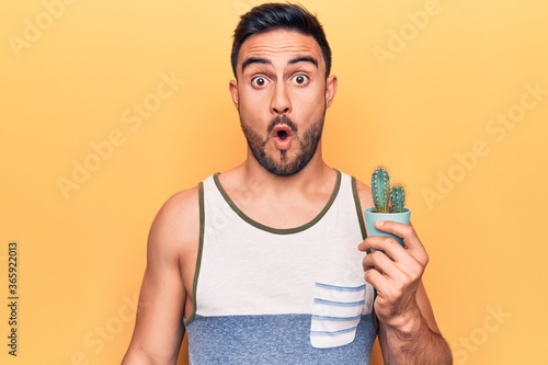Young handsome man with beard wearing sleeveless t-shirt holding cactus plant pot scared and amazed with open mouth for surprise, disbelief face