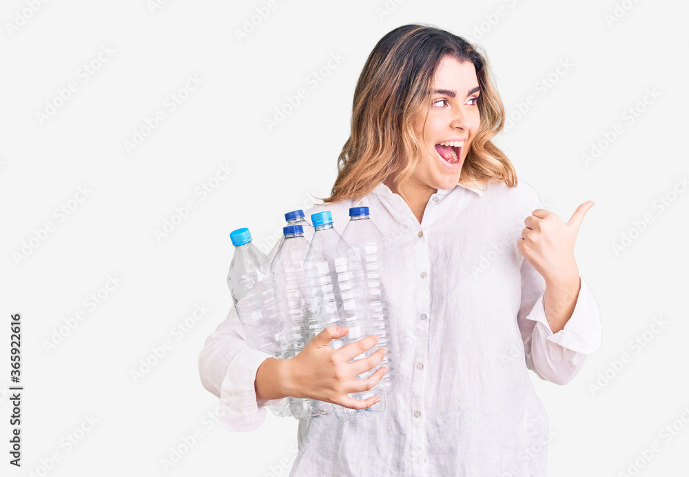 Young caucasian woman holding recycling plastic bottles pointing thumb up to the side smiling happy with open mouth Young caucasian woman holding recycling plastic bottles pointing thumb up to the side smiling happy with open mouth