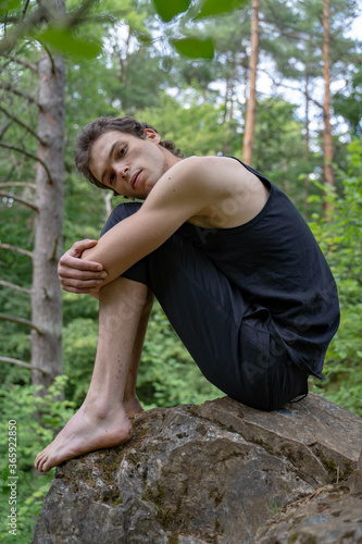 Side view portrait of thoughtful man hugging knees while sitting on rock in forest