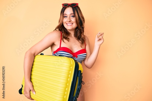 Young beautiful caucasian woman wearing bikini and holding suitcase going on a trip smiling happy pointing with hand and finger to the side