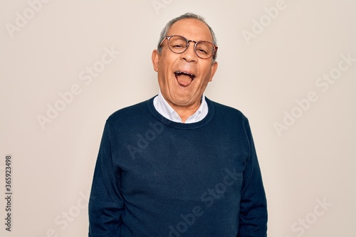 Senior handsome grey-haired man wearing sweater and glasses over isolated white background sticking tongue out happy with funny expression. Emotion concept. © Krakenimages.com