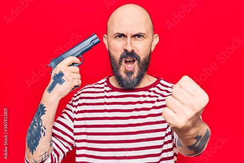Young handsome man holding gun annoyed and frustrated shouting with anger, yelling crazy with anger and hand raised