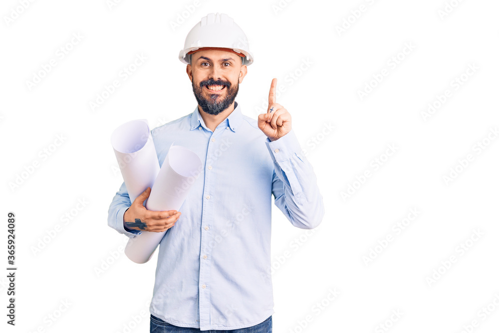 Young handsome man wearing hardhat holding paper blueprints surprised with an idea or question pointing finger with happy face, number one