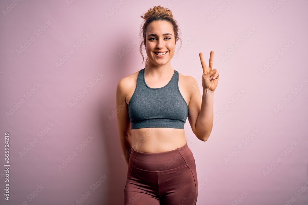 Young beautiful blonde sportswoman doing sport wearing sportswear over pink background showing and pointing up with fingers number two while smiling confident and happy.