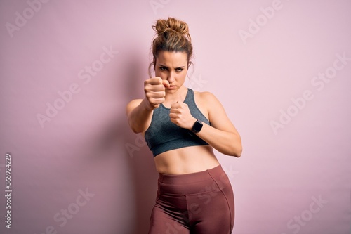 Fototapeta Naklejka Na Ścianę i Meble -  Young beautiful blonde sportswoman doing sport wearing sportswear over pink background Punching fist to fight, aggressive and angry attack, threat and violence