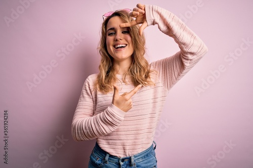 Young beautiful blonde woman wearing casual sweater and sunglasses over pink background smiling making frame with hands and fingers with happy face. Creativity and photography concept.