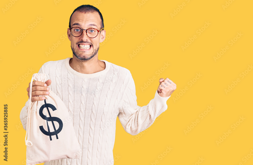 Young handsome man holding money bag with dollar symbol screaming proud, celebrating victory and success very excited with raised arms