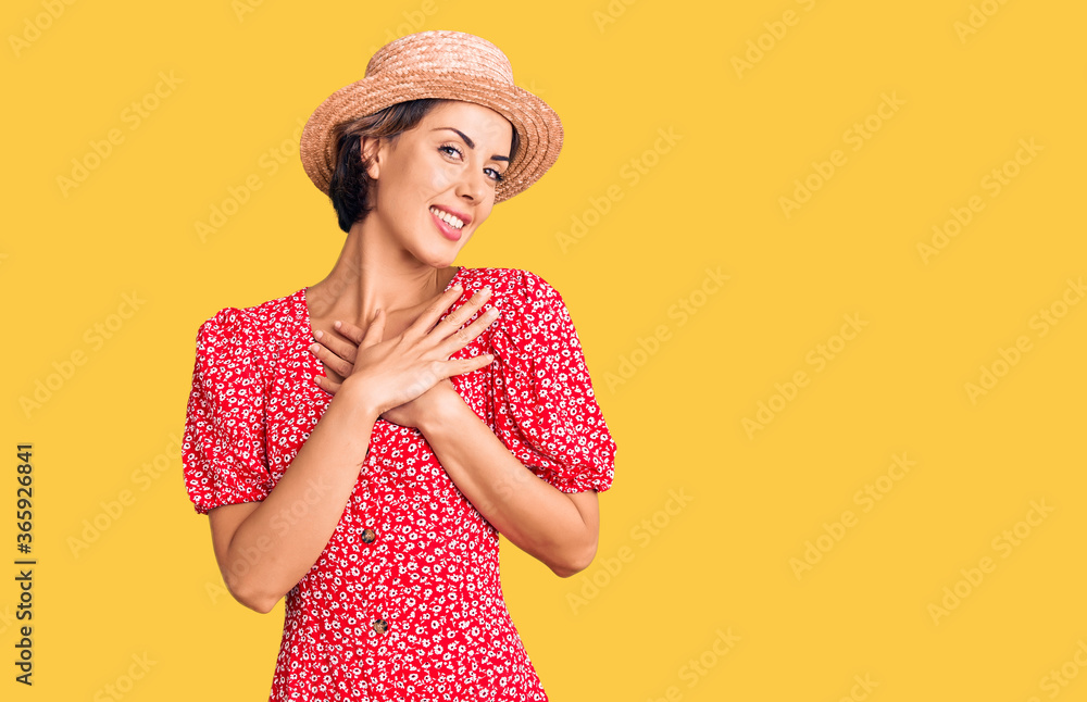 Young beautiful woman wearing summer hat smiling with hands on chest with closed eyes and grateful gesture on face. health concept. Young beautiful woman wearing summer hat smiling with hands on chest with closed eyes and grateful gesture on face. health concept.