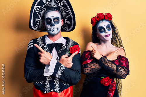 Young couple wearing mexican day of the dead costume over yellow pointing to both sides with fingers, different direction disagree