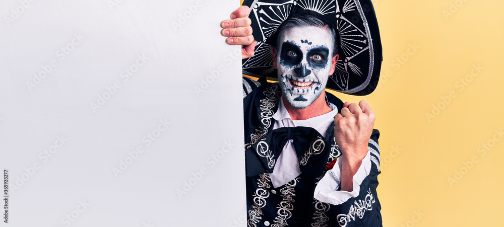 Young man wearing mexican day of the dead costume holding blank empty banner screaming proud, celebrating victory and success very excited with raised arms
