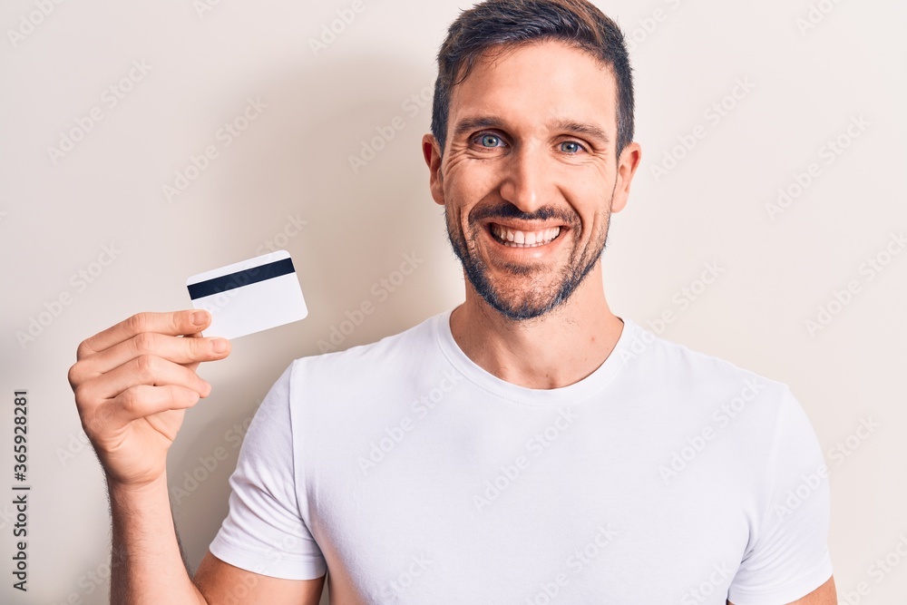 Young handsome customer man holding credit card over isolated white background looking positive and happy standing and smiling with a confident smile showing teeth