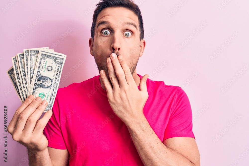 Young handsome man holding bunch of dollars banknotes over isolated pink background covering mouth with hand, shocked and afraid for mistake. Surprised expression Young handsome man holding bunch of dollars banknotes over isolated pink background covering mouth with hand, shocked and afraid for mistake. Surprised expression
