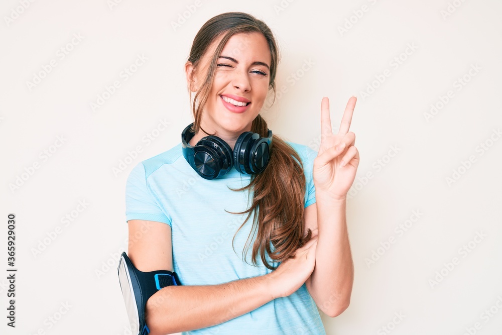Beautiful young caucasian woman wearing gym clothes and using headphones smiling with happy face winking at the camera doing victory sign. number two. Beautiful young caucasian woman wearing gym clothes and using headphones smiling with happy face winking at the camera doing victory sign. number two.